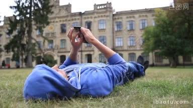 穿着<strong>卫衣</strong>的年轻男学生微笑着躺在大学校园外的草地上，用手机浏览